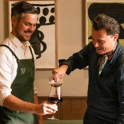 Two men laughing while one pours wine into a glass held by the other, inside a restaurant.