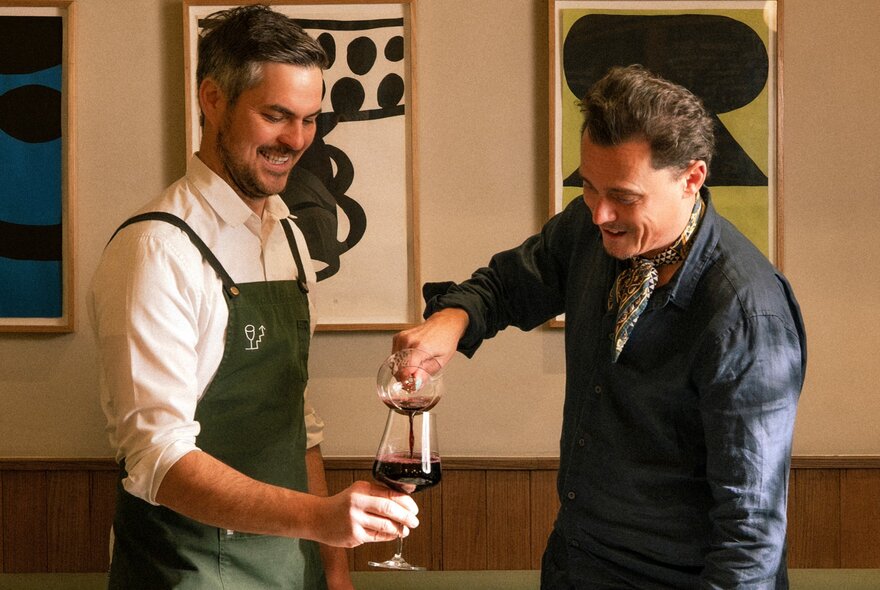 Two men laughing while one pours wine into a glass held by the other, inside a restaurant.