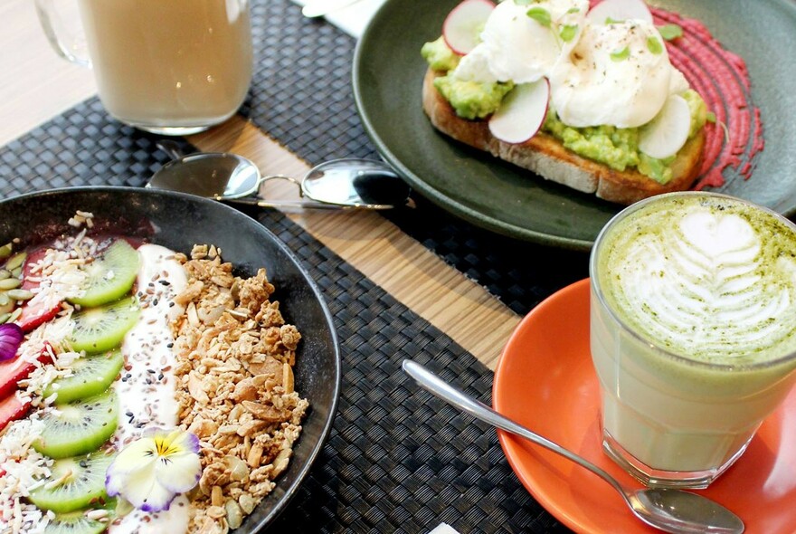 Black placemat with bowl of granola and fruit, another with eggs and smashed avocado, latte to the right.