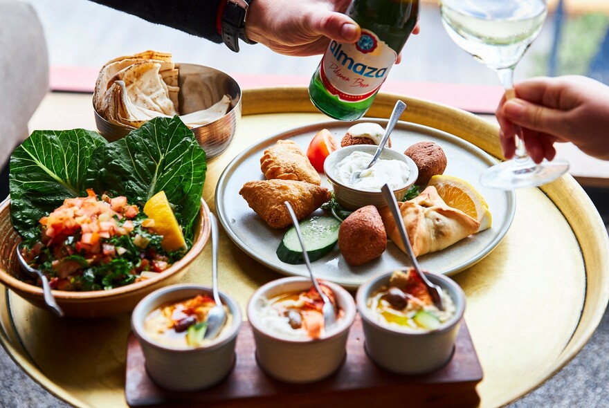 An assortment of snacks and two hands clinking a beer bottle and a wine glass together.