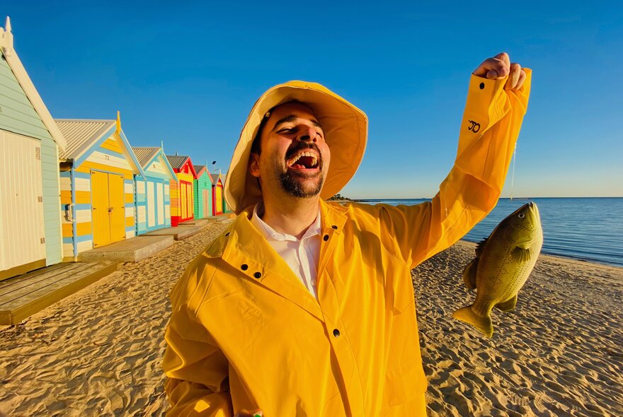 A man wearing a yellow plastic raincoat and hat, laughing while holding a fish that dangles from a line, standing in front of colourful wooden beach bathing boxes at the beach.