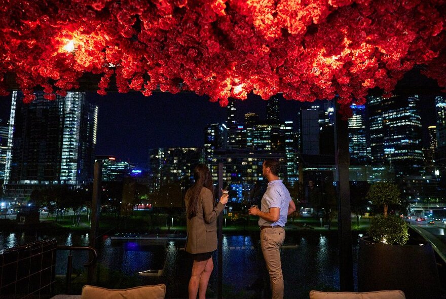 Two people enjoying a cocktail looking out over the city from a rooftop bar with a red floral ceiling decoration.