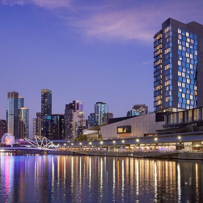 High-rise hotel positioned beside the Yarra River with lights reflected in the water at dusk.
