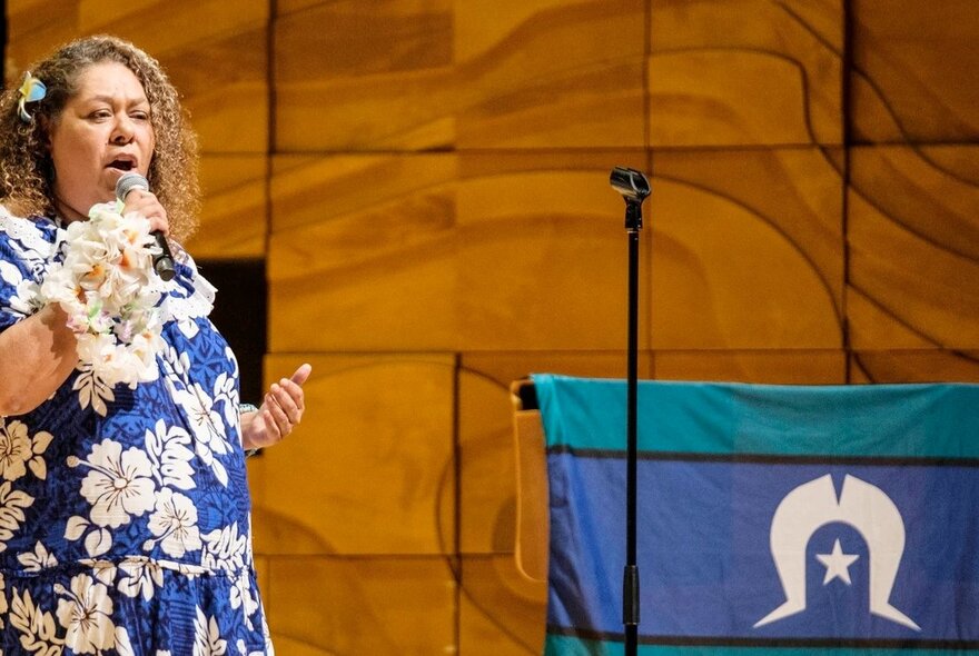 A woman wearing a floral dress singing into a microphone, standing on a stage next to a Torres Strait Island flag.
