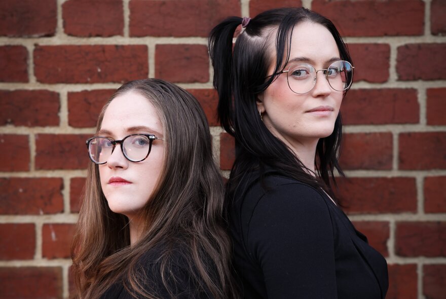 Two women wearing glasses standing back-to-back with a brick wall behind them. 
