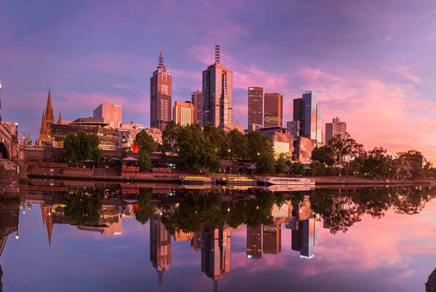 A wide angle shot of the North Bank of Melbourne's Yarra River at dusk, with buildings and trees reflected in the still water.