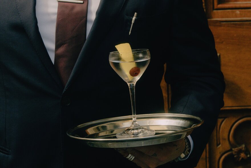 A classic martini garnished with an olive and a broad lemon peel twist, served in a small V-shaped cocktail glass balanced on a silver tray being carried by a waiter.