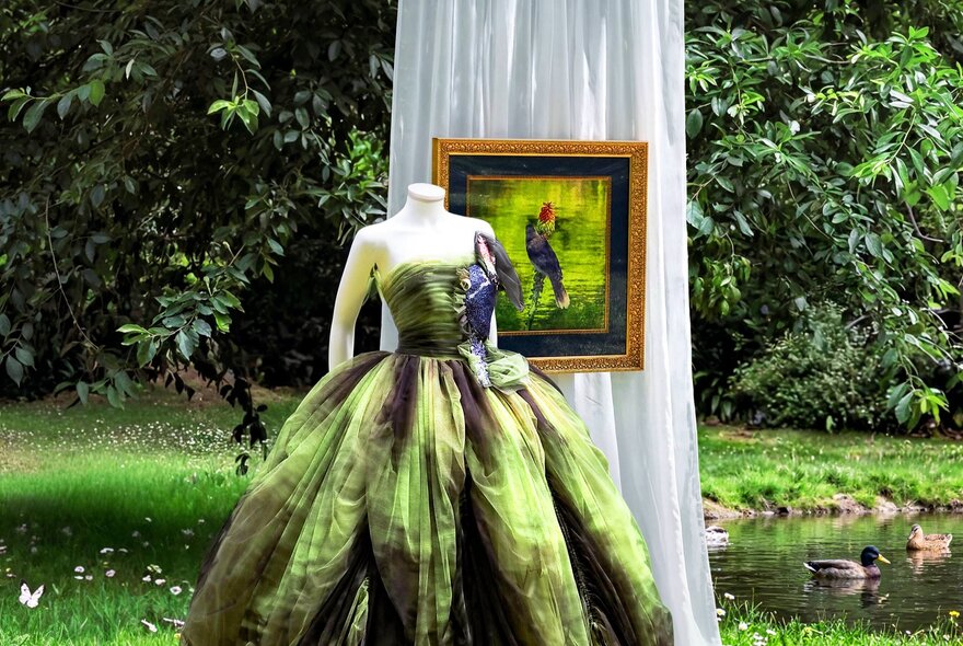 A printed green strapless ballgown on a headless mannequin in a garden with a white draped curtain and a framed picture.