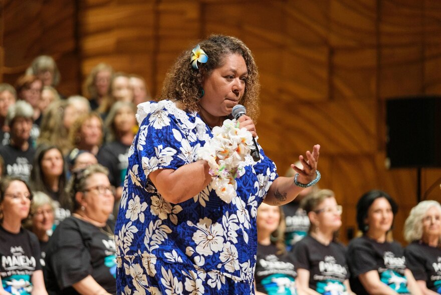 A woman in a floral dress with frangipani in her hair and on her wrist, singing to an audience with choir members in the background.
