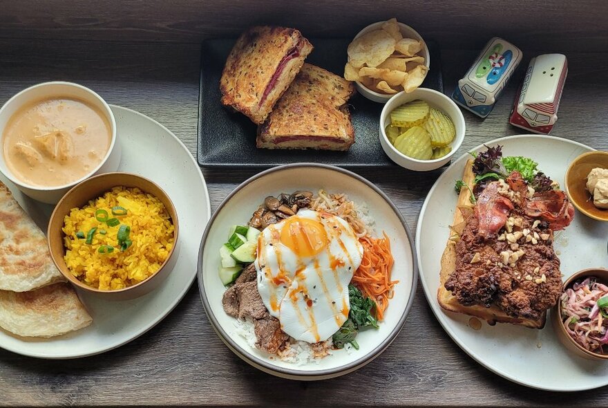 Looking down at a table from above, with plates of food including a fried egg on salad and noodles, a toasted sandwich, rice, laksa and roti bread.