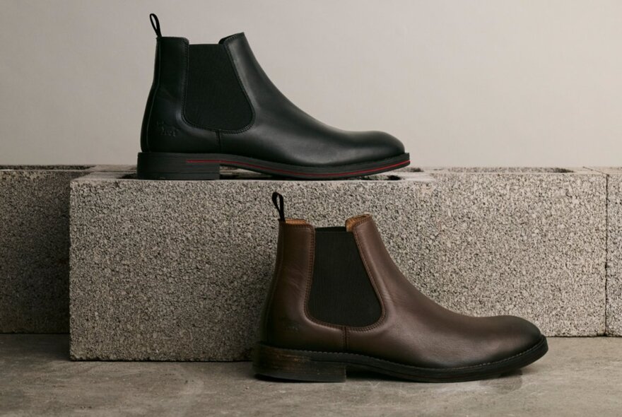 Two Chelsea boots in profile, one black on a stone block and one brown on a floor. 