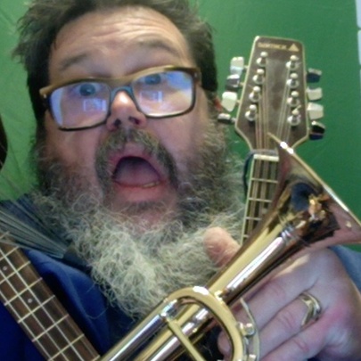 A bearded man with glasses looking aghast with a ukulele, guitar and trumpet draped over him. 