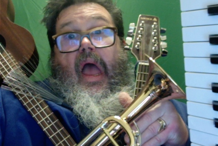 A bearded man with glasses looking aghast with a ukulele, guitar and trumpet draped over him. 