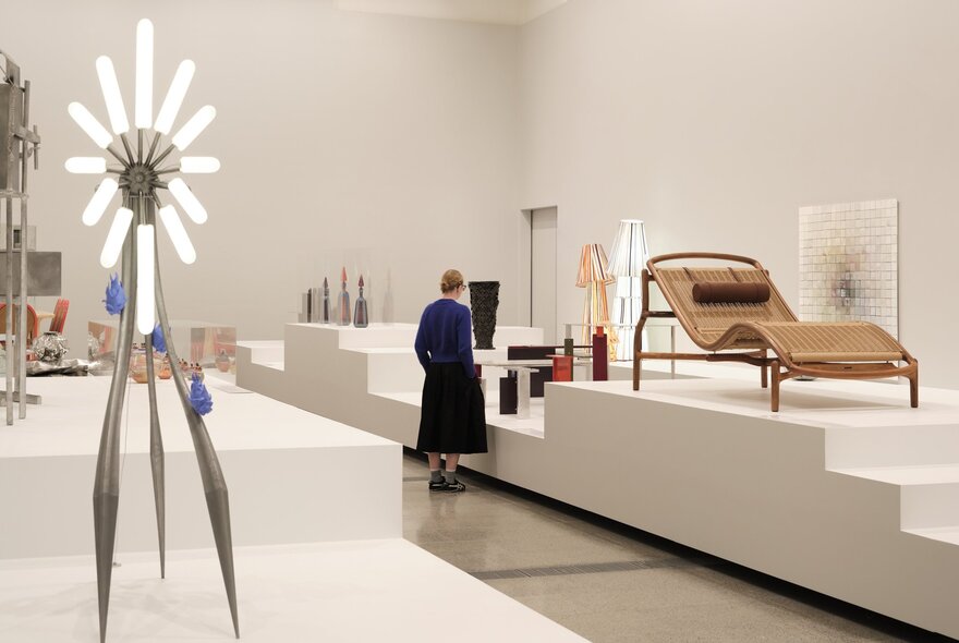 Creative design artworks displayed in a white gallery setting, with some artworks hanging on the wall and others on a white dais; a person viewing the collection.