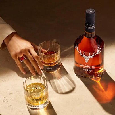 A hand reaching into the frame towards a short glass of whiskey, with a bottle of Dalmore whiskey alongside.