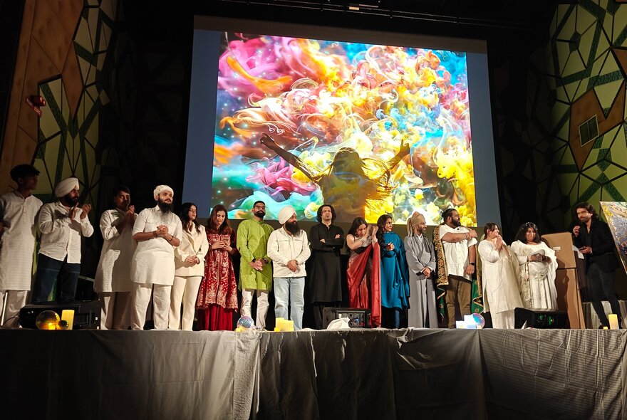 An event on a stage, featuring a diverse group of individuals dressed in varied attire standing in a row, with a large screen behind them.