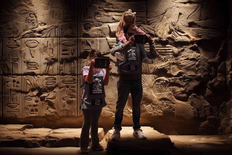 A father with his two daughters, one on his shoulders, each looking at a digital device in front of a wall decorated with Ancient Egyptian-style wall carvings.