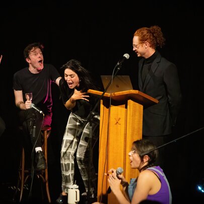 Six comedians on stage all laughing hysterically, one standing behind a wooden podium.