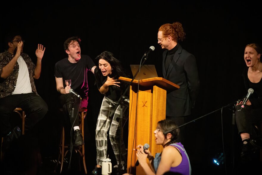 Six comedians on stage all laughing hysterically, one standing behind a wooden podium.