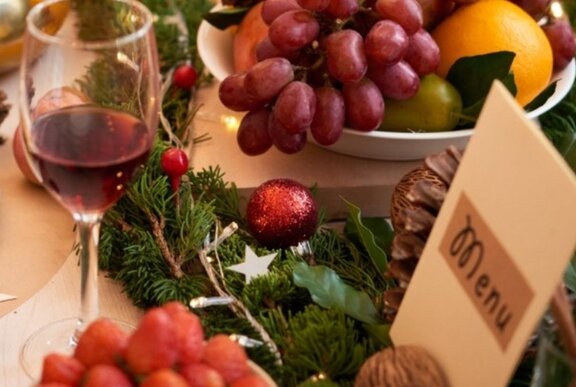 A glass of red wine alonside a festive wreath with red baubles and silver stars, a menu card and a bowl of fruit.