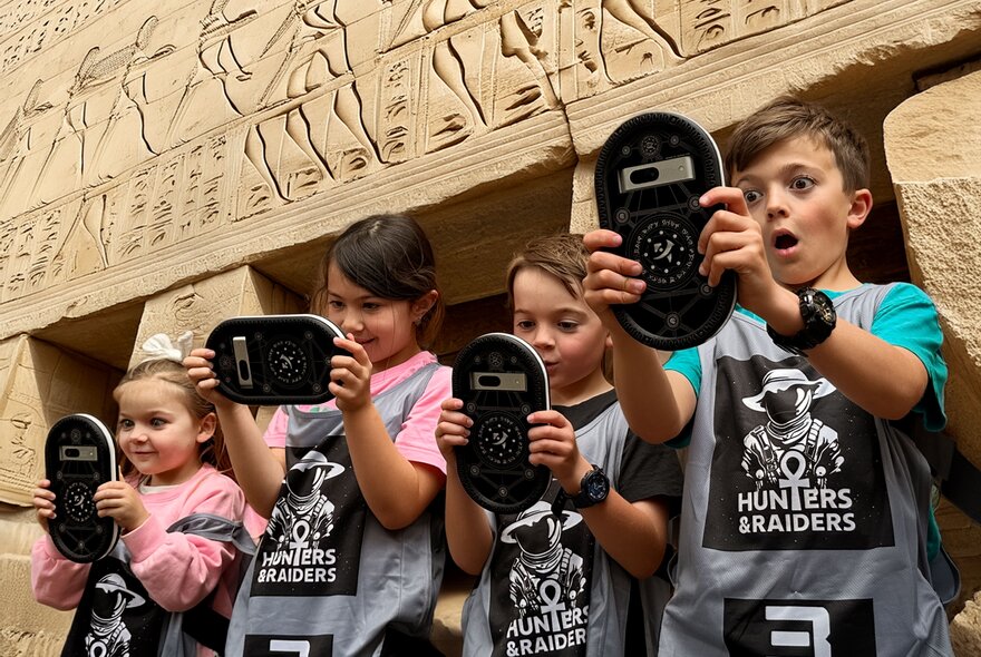 Four children holding digital devices while standing in front of an Anciant Egyptian-style wall carving.