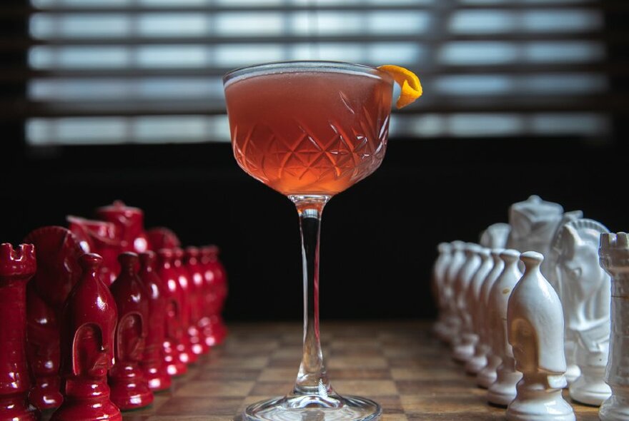 A chess board with red and white pieces lined up to play and a berry-coloured cocktail in the middle of the board.