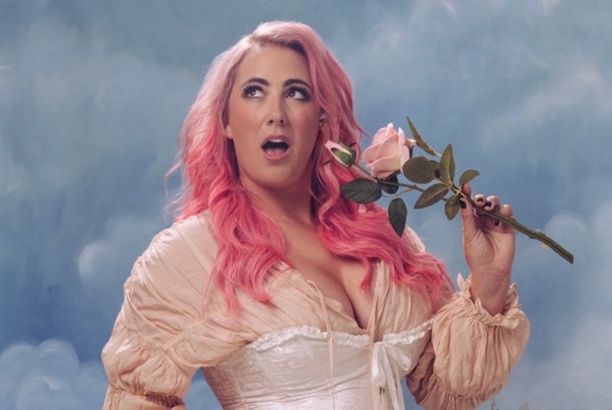 Comedian with long pink hair wearing a bustier and lace top, holding a rose in front of a cloudy blue sky.