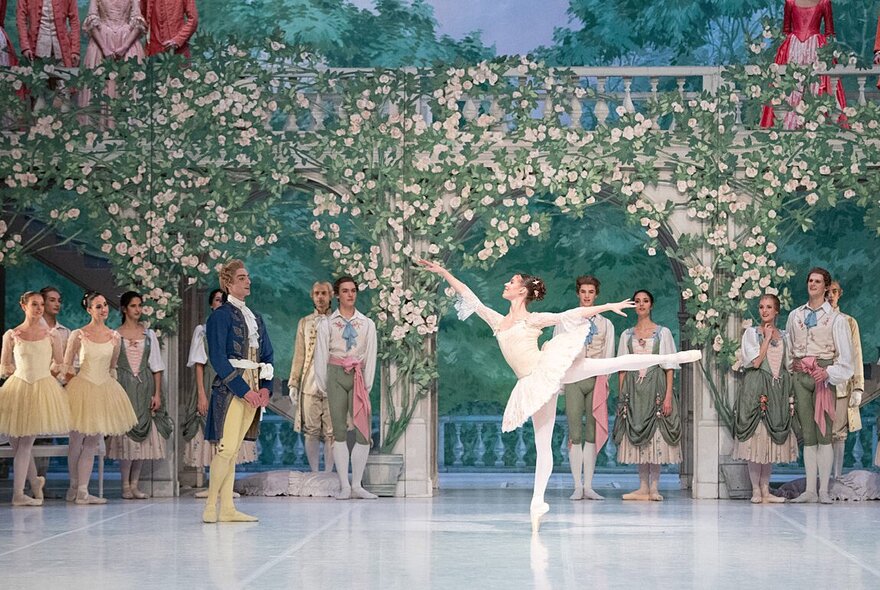 A ballerina performs a pose with one leg in the air as the troupe looks on.