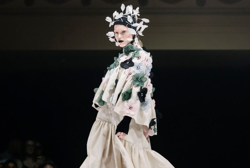 A model wearing a layered ensemble with floral embellishments and an elaborate headpiece on a runway.