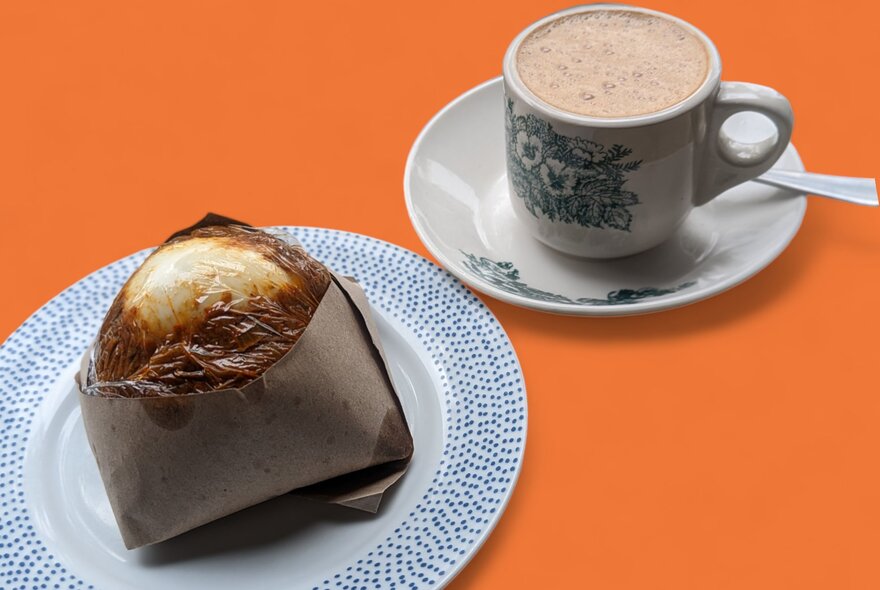 Looking at an orange table with a cup of coffee and a pastry wrapped in a brown paper pocket
