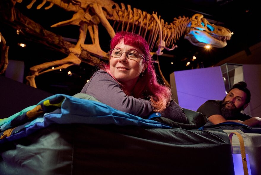 A woman with bright red hair smiling from a bed underneath a large dinosaur skeleton.