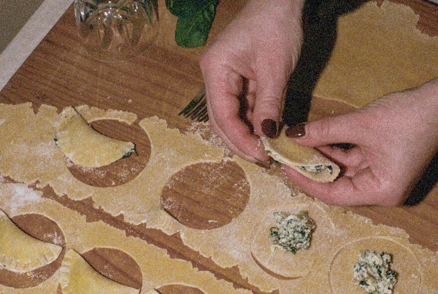 A pair of hands folding a spinach and ricotta mixture into a half-moon shaped agnolotti on a wooden kitchen table.