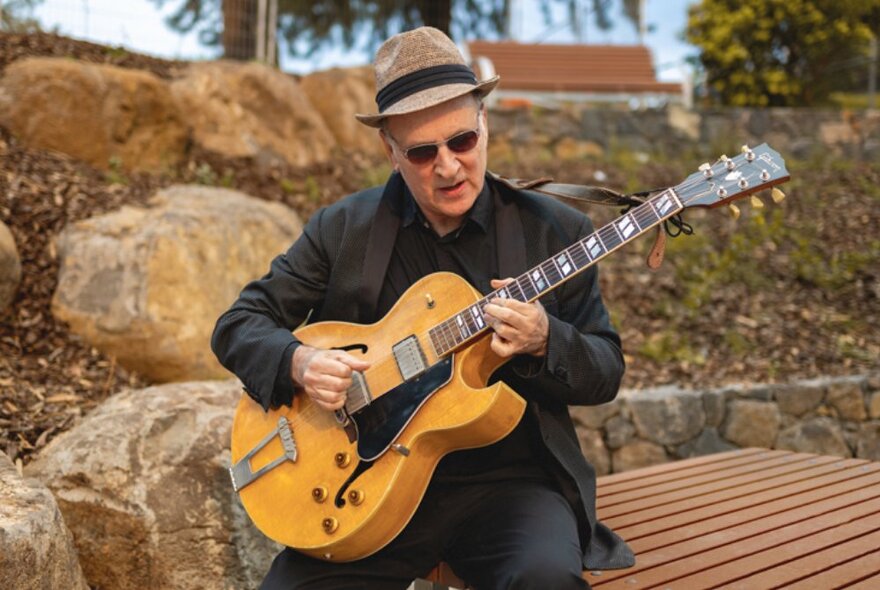 Man wearing a hat seated on an outside bench playing a guitar.