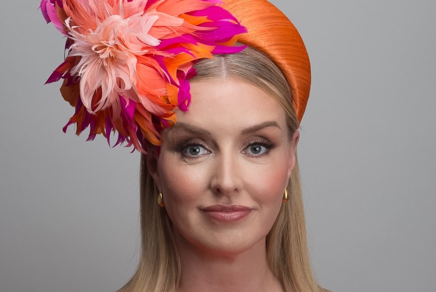 Headshot of a woman modelling a bright orange and fuchsia coloured floral fascinator. 