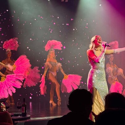 A cabaret singer on stage with dancers with fans, wearing a glittering gown with arm outstretched. 