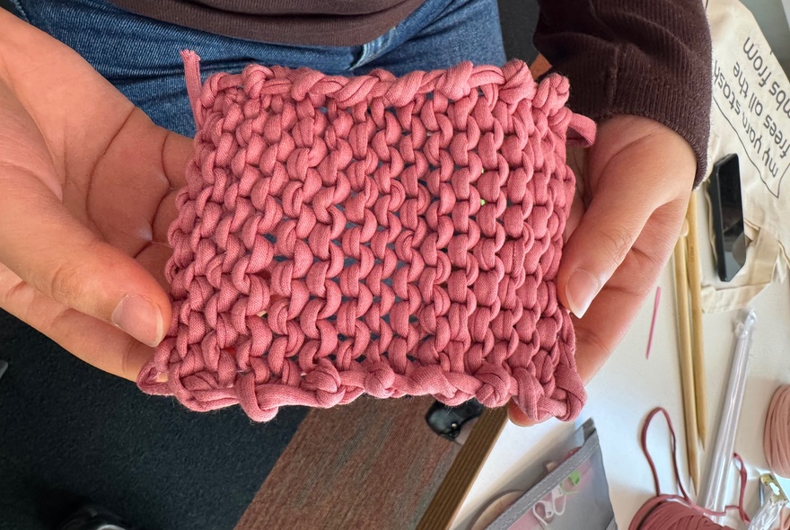A close-up view of two hands holding a small, square fabric swatch knitted with thick pink yarn in a bumpy stitch pattern, with knitting needles visible in the background.