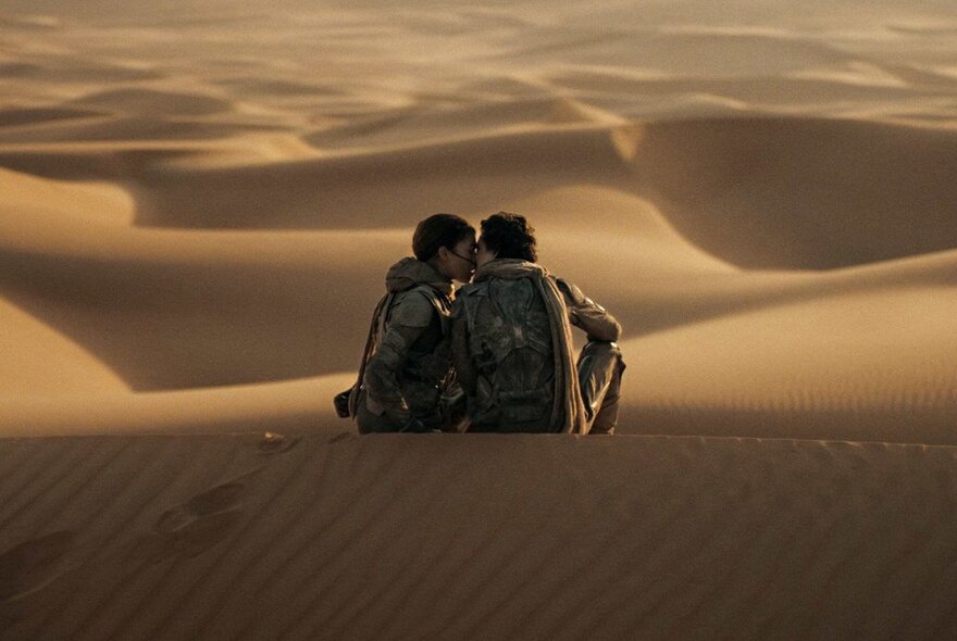 A still from the movie Dune: Part Two showing two people sitting on sandy dunes next to each other kissing.