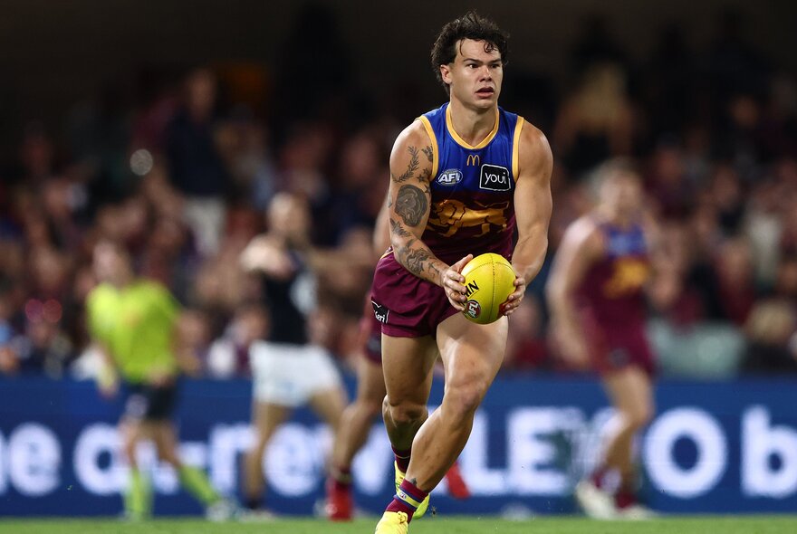 A Brisbane Lions AFL player running with the football as if about to kick it, blurred players, umpires and footy fans in the stadium behind him.
