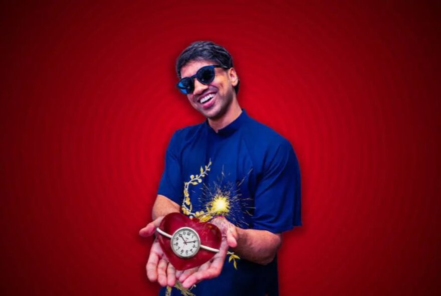 Smiling man in a blue shirt and sunglasses, holding in his open hands a red, heart-shaped object resembling a bomb with a clock face and a fuse sparking.