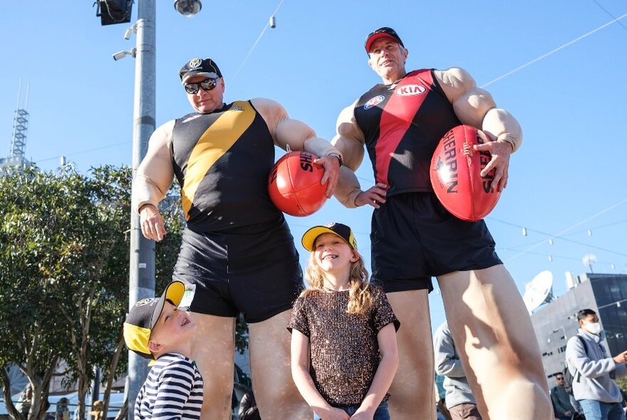 Two young children posing underneath two footballers wearing oversized puffy suits; photo taken from the ground and looking up.