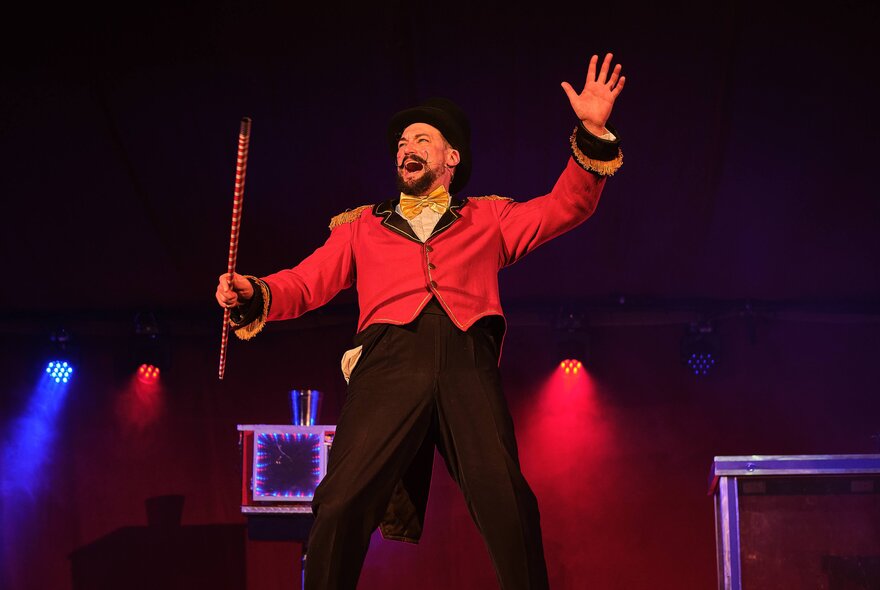 A male performer dressed in a red ringmaster-style jacket and black trousers on a stage with blue and red lighting, holding a stick and gesturing with his other hand during a circus and magic show.
