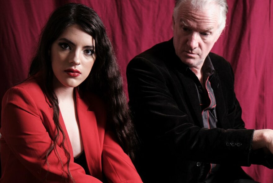 Performers, Mick Harvey and Amanda Acevedo, seated next to each other in front of a red stage curtain.