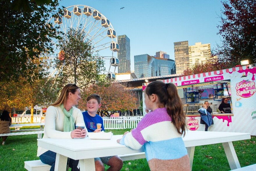 A family seated at an outdoor table in a grassy park, eating takeaway food from the food van parked behind them, the Skyline Ferris wheel in the background.