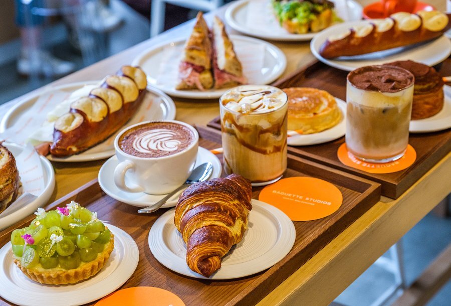 A table set with cafe dishes including coffees, pastries and pretzel baguettes.