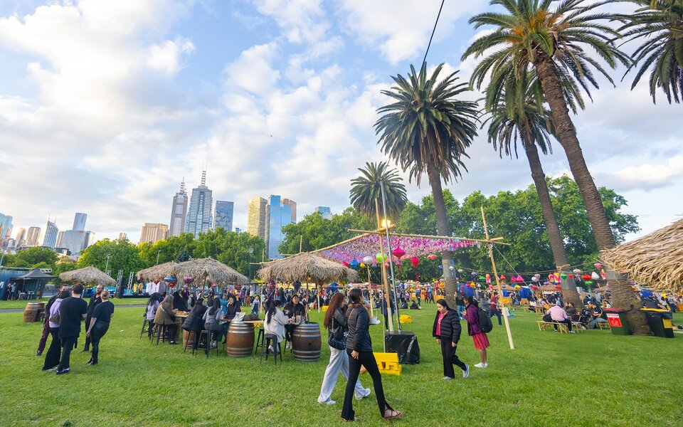 Crowds enjoying an outdoor festival in a green space next to the city, with lanterns and tiki huts.