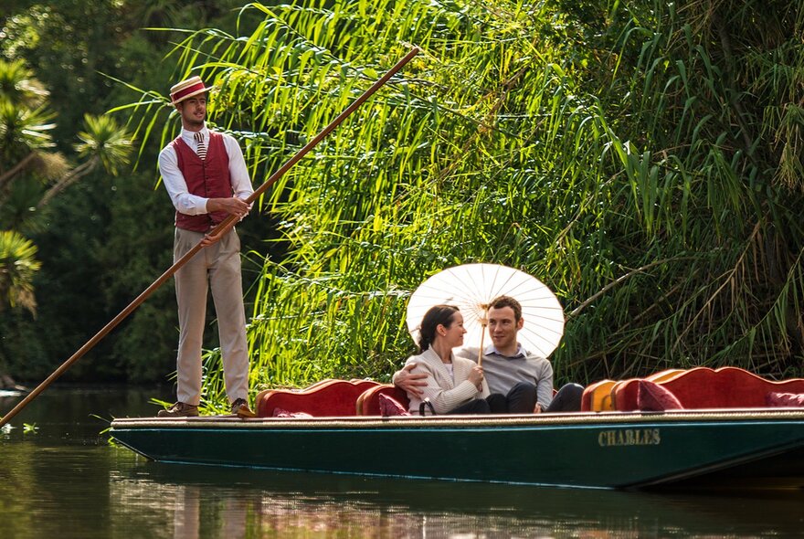 A couple in a small boat under a parasol while a man in a vest and hat steers the boat with a long stick.