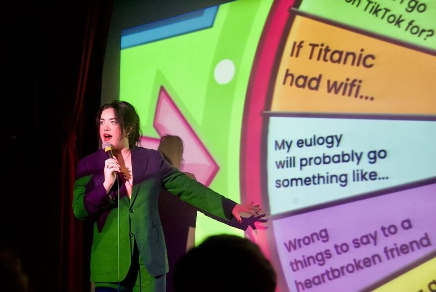 A comedian on stage in front of a projected spinning wheel that has prompt phrases printed in the segments.