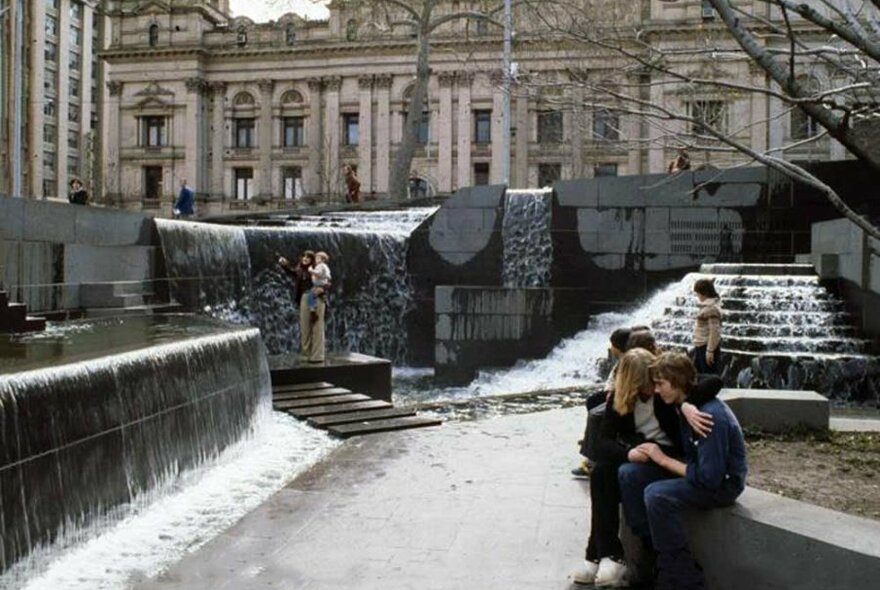 Two people sitting next to fountains in a city square.