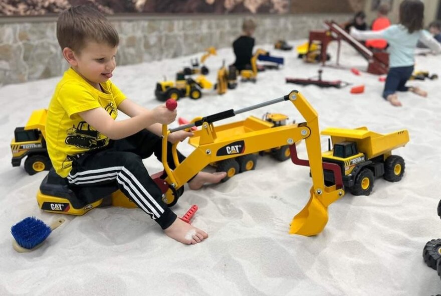 A small boy plays with a ride-on digger in a large sandpit. 