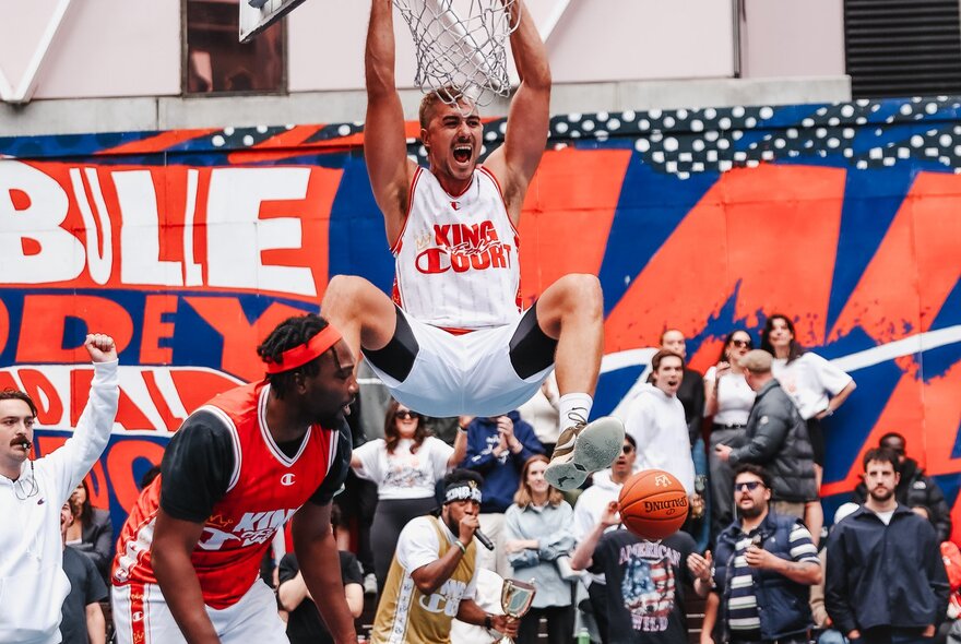 A man hanging off a basketball hoop after a slam dunk at a street basketball court in front of an audience.